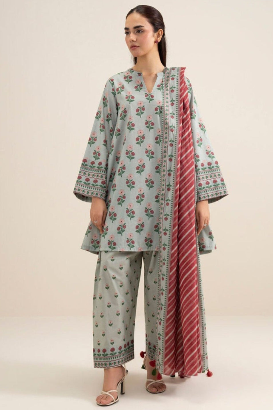 A model wearing a three-piece printed khaddar suit from the brand SAPPIRE, featuring a light sage green kurta and trousers with red floral patterns, paired with a red and white striped dupatta. The image is a product information card with text details, care instructions, and a barcode on a white background.