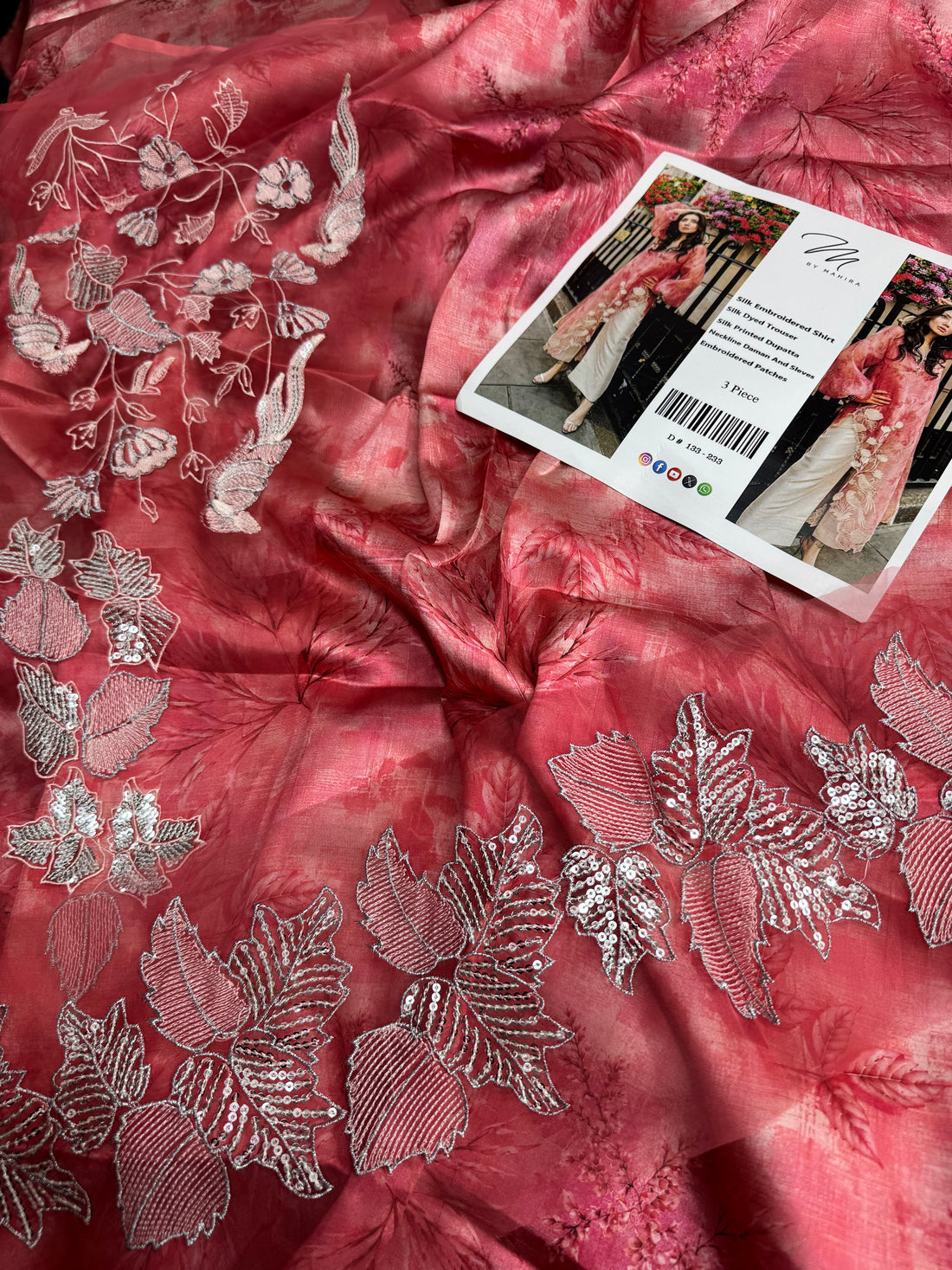 A woman models a pink floral printed long kurta with sheer balloon sleeves and white floral embroidery along the hem, paired with cream wide-leg trousers, standing outdoors in front of a black metal fence.