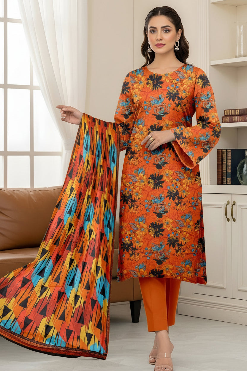A woman modeling a Kalkaram Bana Dora Lawn/Borderline set, featuring a bright orange kurta with black floral patterns, matching orange pants, and a coordinating printed dupatta, set against an indoor home background.