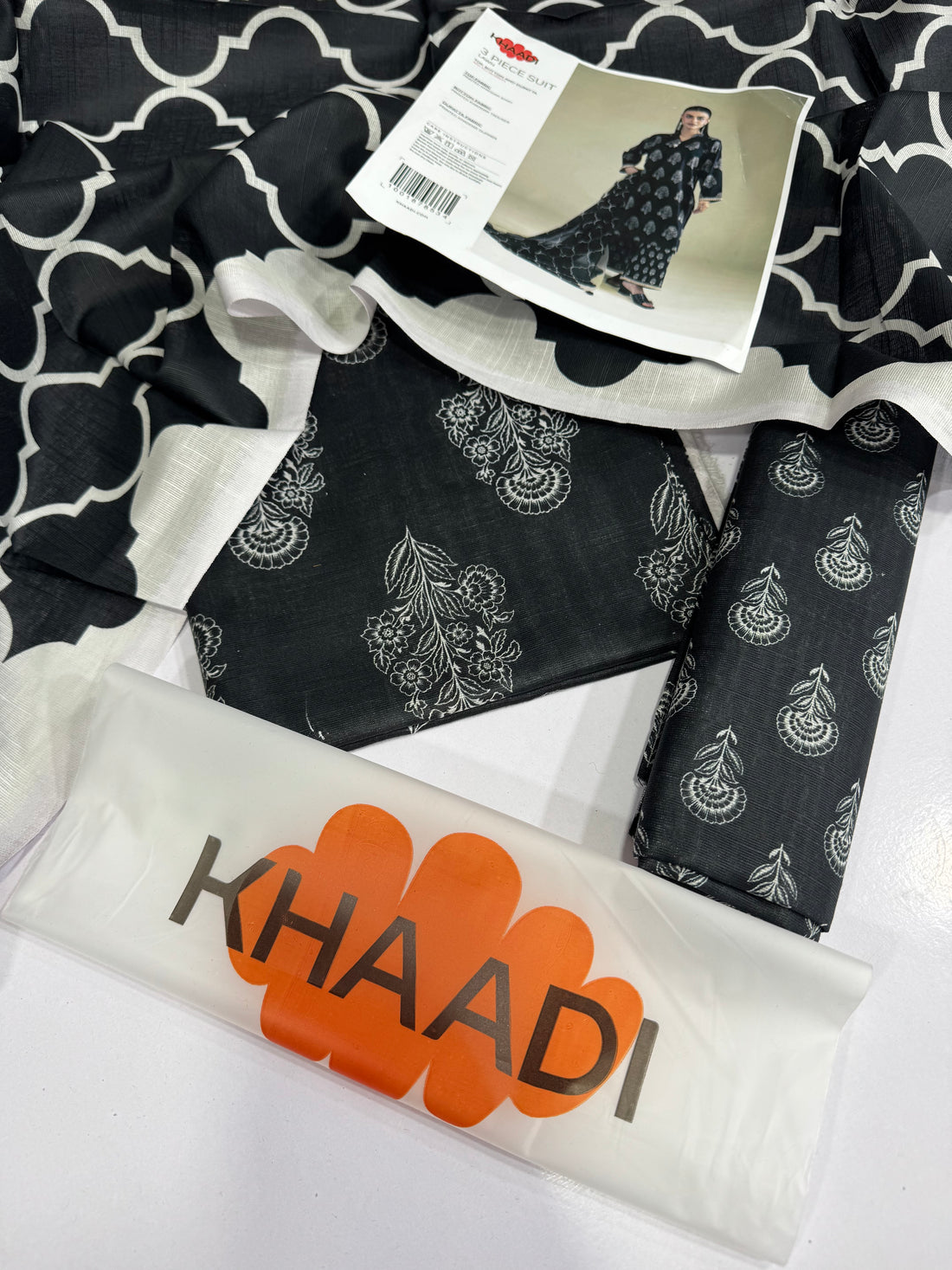 A model wearing a black three-piece suit with white printed motifs, shown against a light beige studio background. The left side of the image features a white panel with product details, care instructions, and a barcode under the brand name KHAADI.
