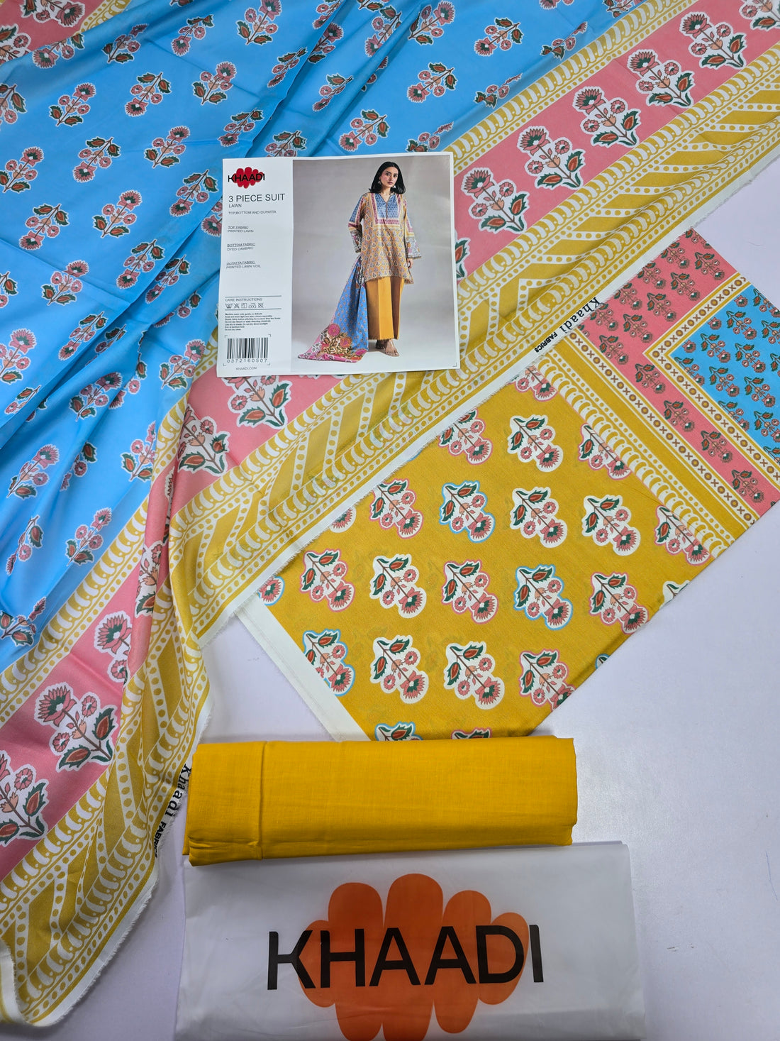 A 3-piece printed lawn suit by Khaadi, featuring a tunic with multicolor floral patterns, mustard yellow pants, and a matching blue dupatta with pink and orange floral motifs, shown on a model against a light gray background with product details on the left.