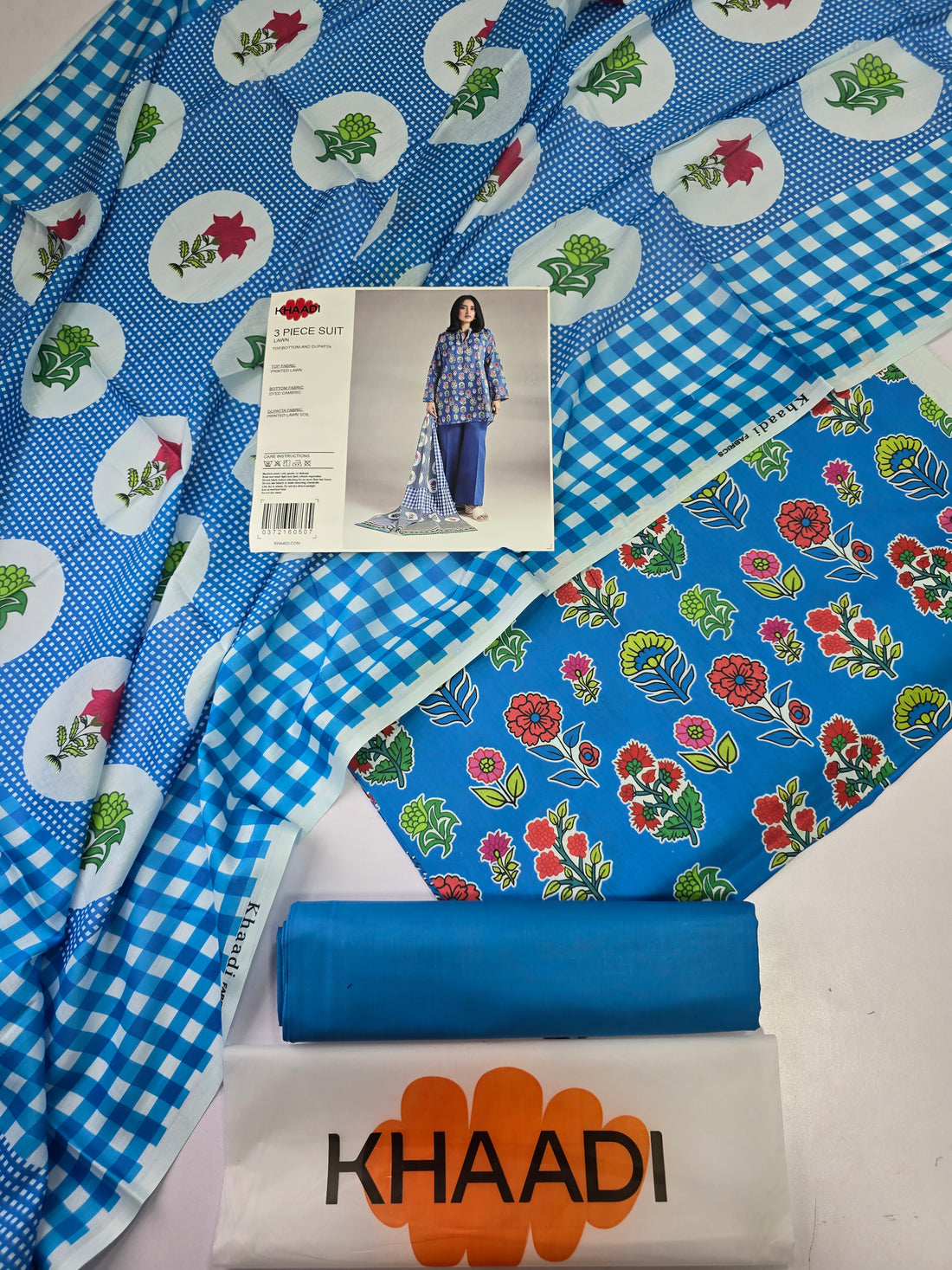 A 3-piece women's suit set from KHAADI, featuring a blue printed kurta, solid blue pants, and a blue-and-white gingham dupatta with colorful paisley borders, shown on a model against a light gray studio background with product details listed on the left.