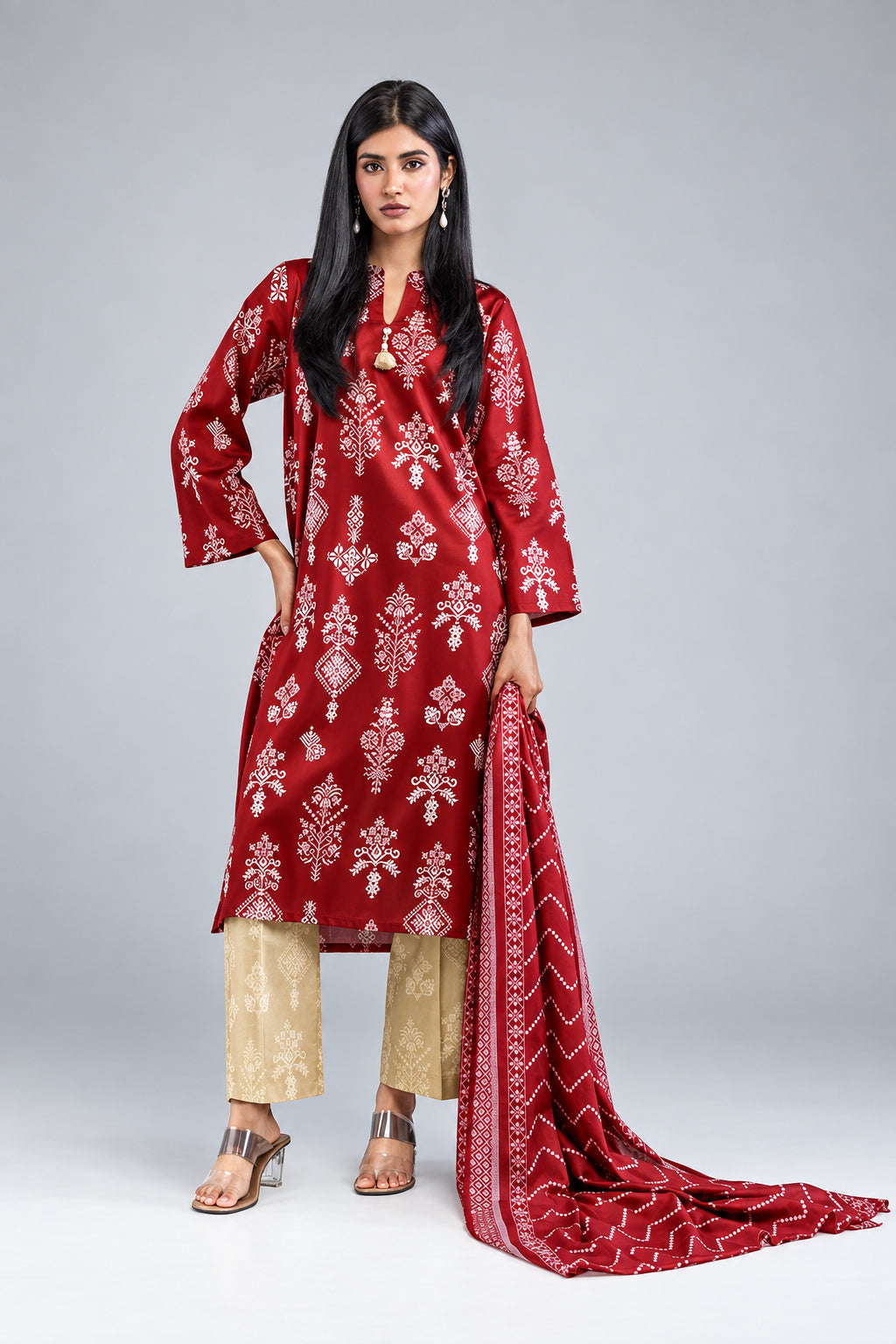 A model wearing a Khaadi 3-piece suit in lawn, featuring a red printed kurta, beige pants, and a matching red dupatta, set against a light beige studio background with product details on the left.