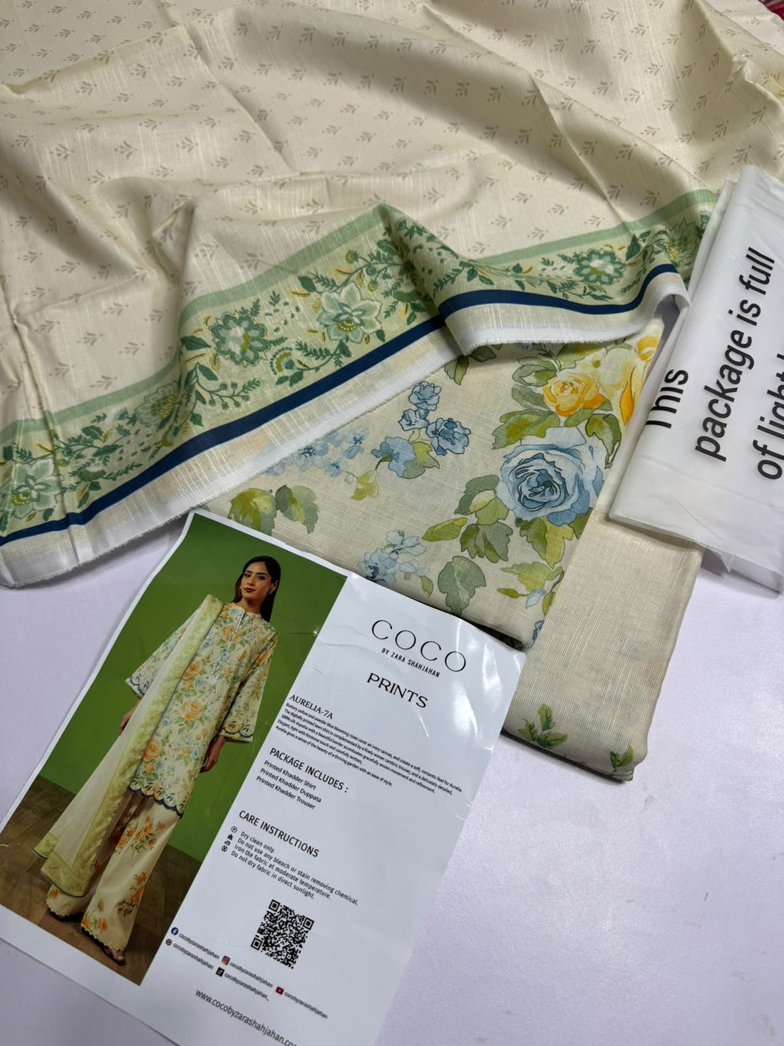 A model wearing a printed traditional outfit stands against a green studio backdrop, with printed text panels on the right showing the brand name COCO BY ZARA SHAHJAHAN, product name AURELIA-7A, package details, and care instructions.