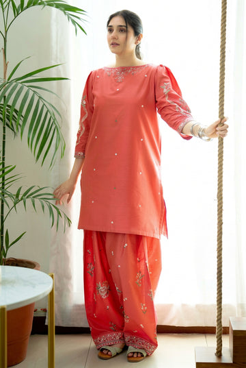A woman modeling a coral pink embroidered Dhanak two-piece suit from the URGPEET Winter Collection, consisting of a long kurta and wide-leg trousers, standing next to a rope prop.