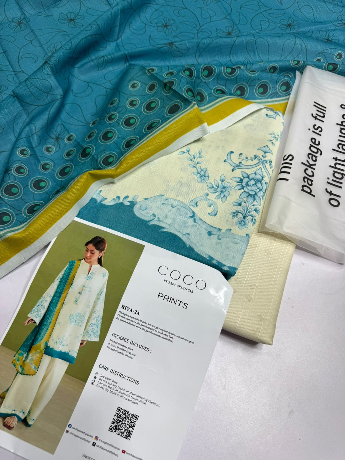 A model wearing a three-piece women's salwar kameez suit from Coco by Zara Shahahan, featuring a cream-colored printed kameez, matching trousers, and a teal and yellow dupatta, set against a split background with product details and text.