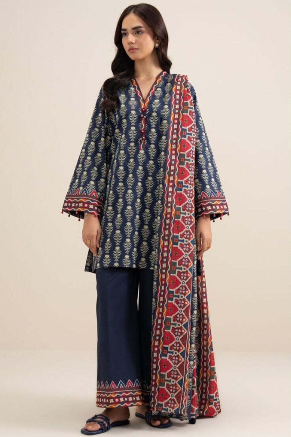 A model wearing a three-piece printed khaddar suit by Sapphire, featuring a long kurta, trousers, and a top, displayed on a clean white catalog card with product details and care instructions.