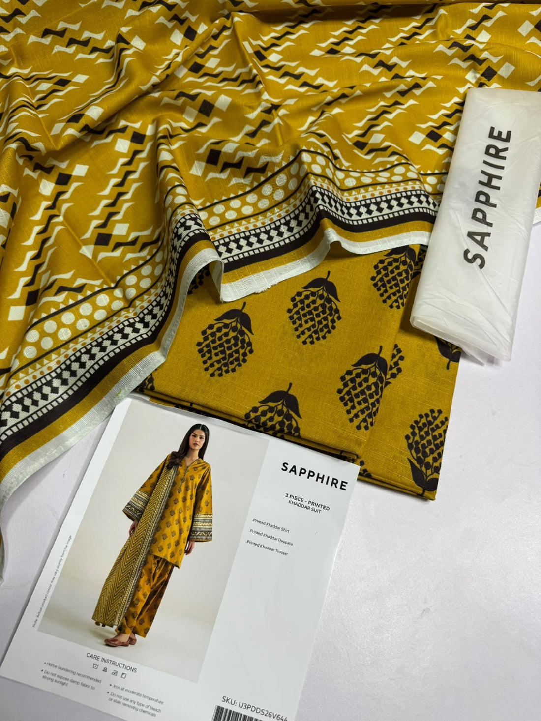 A model wearing a three-piece printed khaddar suit from the brand SAPPHIRE, featuring a mustard yellow kurta with black and gold patterns, matching trousers, and a dupatta, set against a clean white background with product details and care instructions.