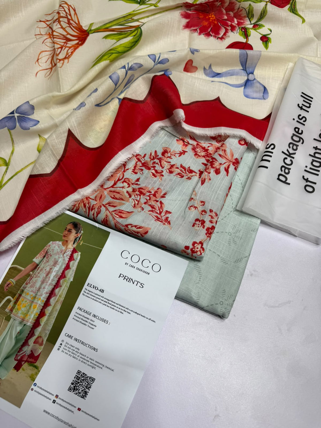 A fashion catalog page showing a model wearing a printed three-piece salwar kameez set in Elyo-4B by Zara Shahrihan, alongside product details, care instructions, and a QR code on a split green-and-white background.