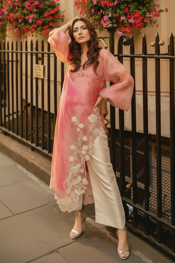 A woman models a pink floral printed long kurta with sheer balloon sleeves and white floral embroidery along the hem, paired with cream wide-leg trousers, standing outdoors in front of a black metal fence.