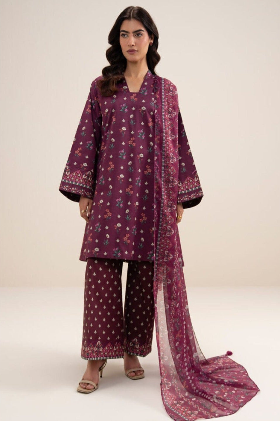A model wearing a three-piece printed khaddar suit from Sapphire, featuring a dusty purple kurta, matching trousers, and a dupatta with small yellow and white floral patterns, set against a plain light beige background with product details on the right.