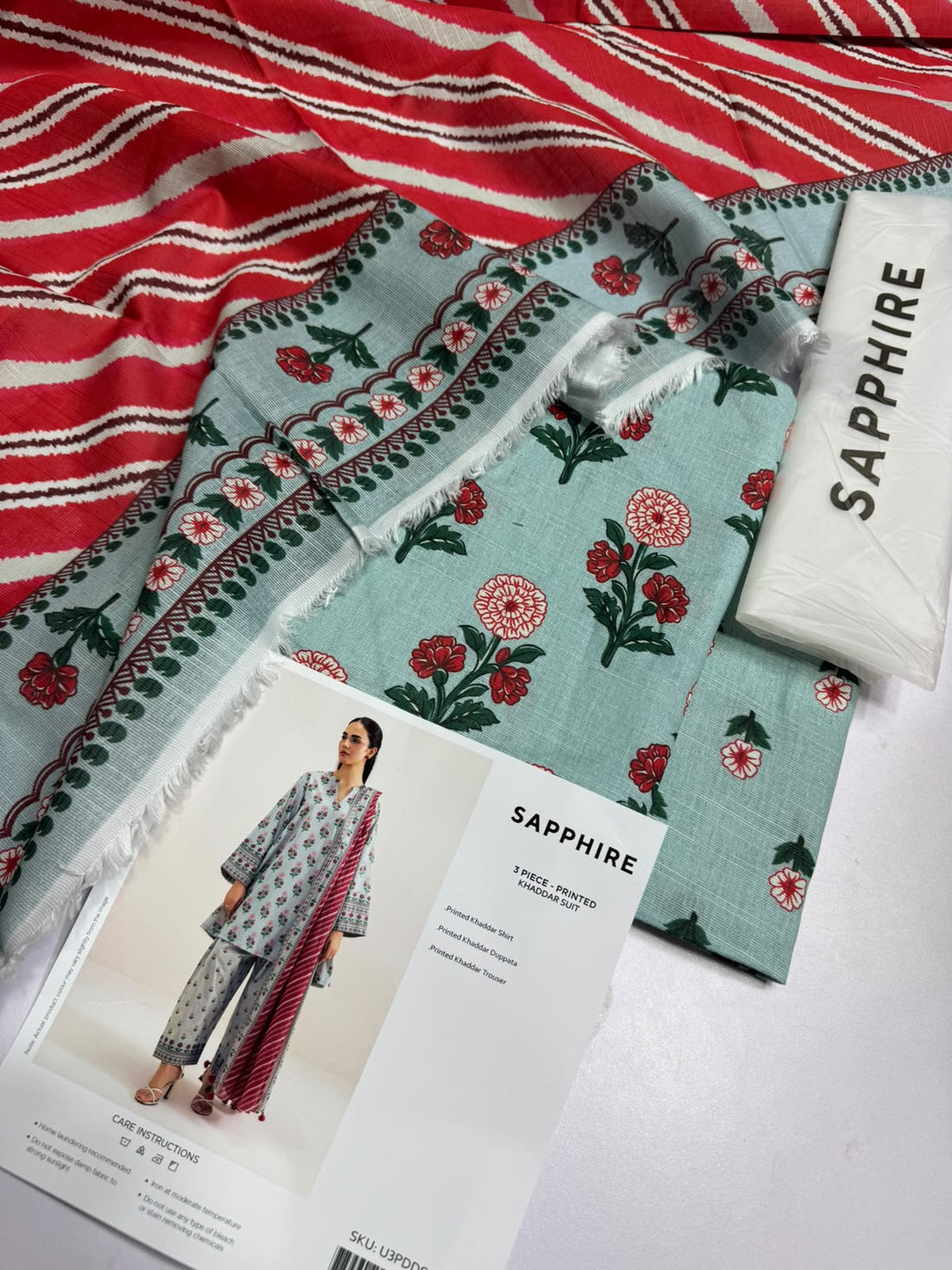 A model wearing a three-piece printed khaddar suit from the brand SAPPIRE, featuring a light sage green kurta and trousers with red floral patterns, paired with a red and white striped dupatta. The image is a product information card with text details, care instructions, and a barcode on a white background.