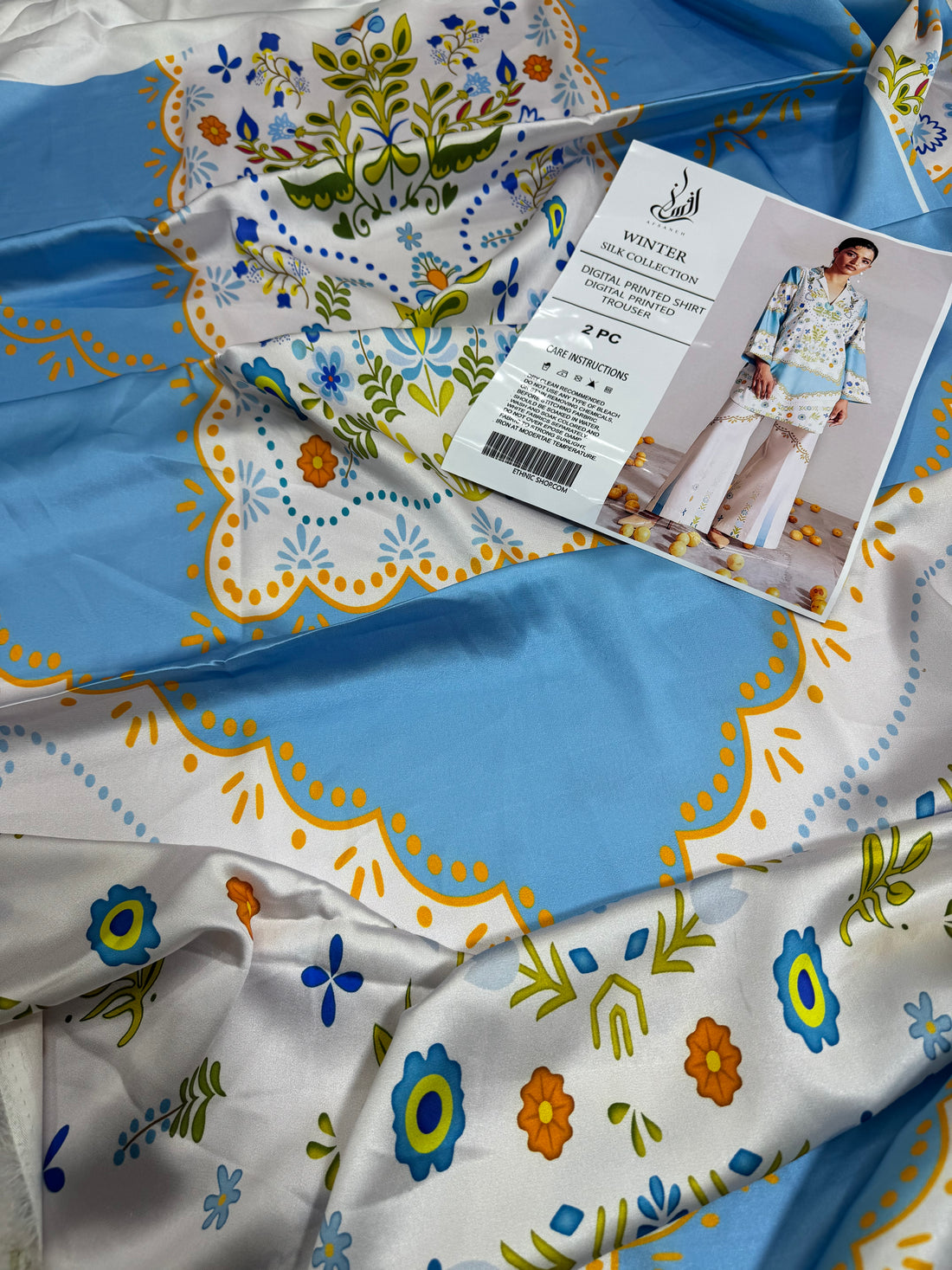 A model wearing a light blue and white printed two-piece kurta and trouser set from Ethnic Shop, shown on a split graphic background with product details and care instructions on the left.