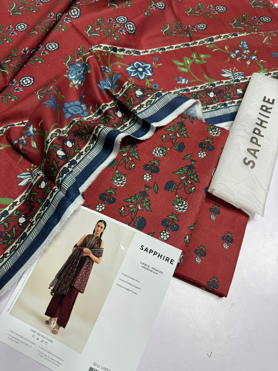 A model wears a three-piece printed khaddar suit from Sapphire, featuring a dark red printed shirt, matching trousers, and a dupatta, displayed on a clean beige background with product details and care instructions.