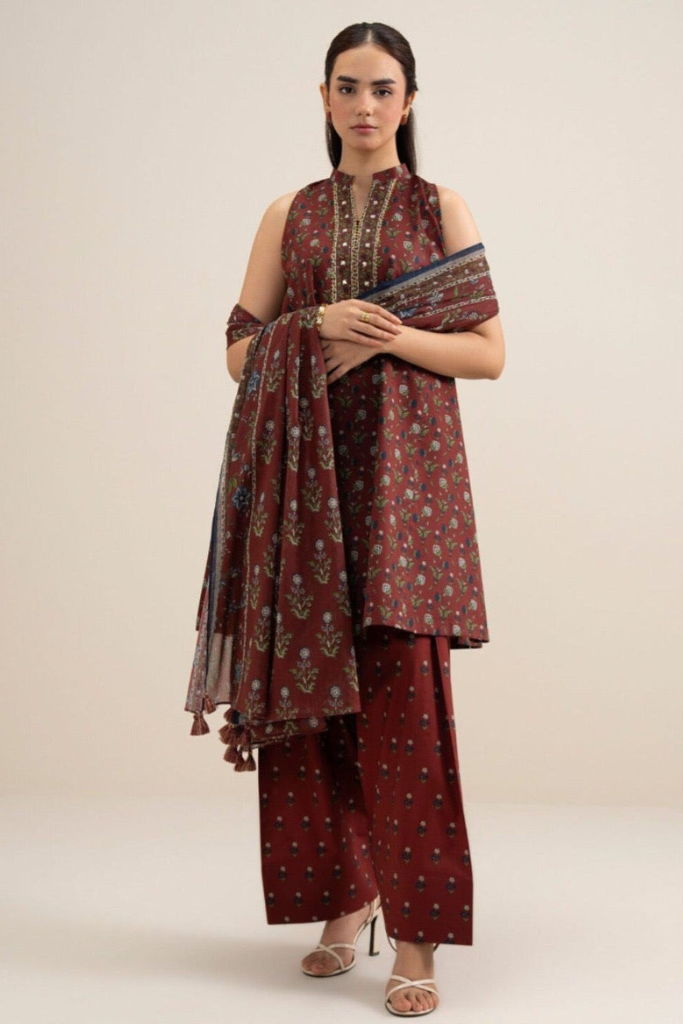 A model wears a three-piece printed khaddar suit from Sapphire, featuring a dark red printed shirt, matching trousers, and a dupatta, displayed on a clean beige background with product details and care instructions.