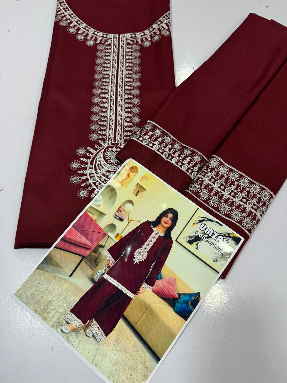 A woman modeling a deep maroon three-piece salwar kameez suit with silver mirror work embroidery on the neckline, sleeve cuffs, and pant hems, standing in a modern living room setting.