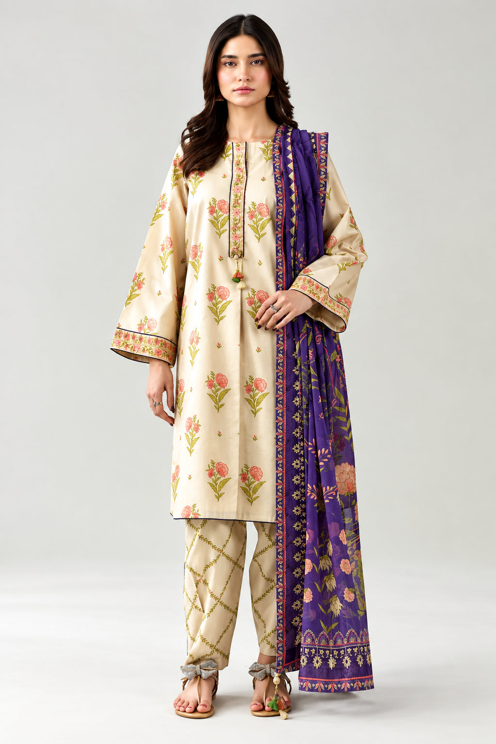 A woman modeling a three-piece Nishat Unstitched digital printed khadgar set. The set includes a beige kurta with floral prints, matching beige trousers, and a purple printed dupatta.