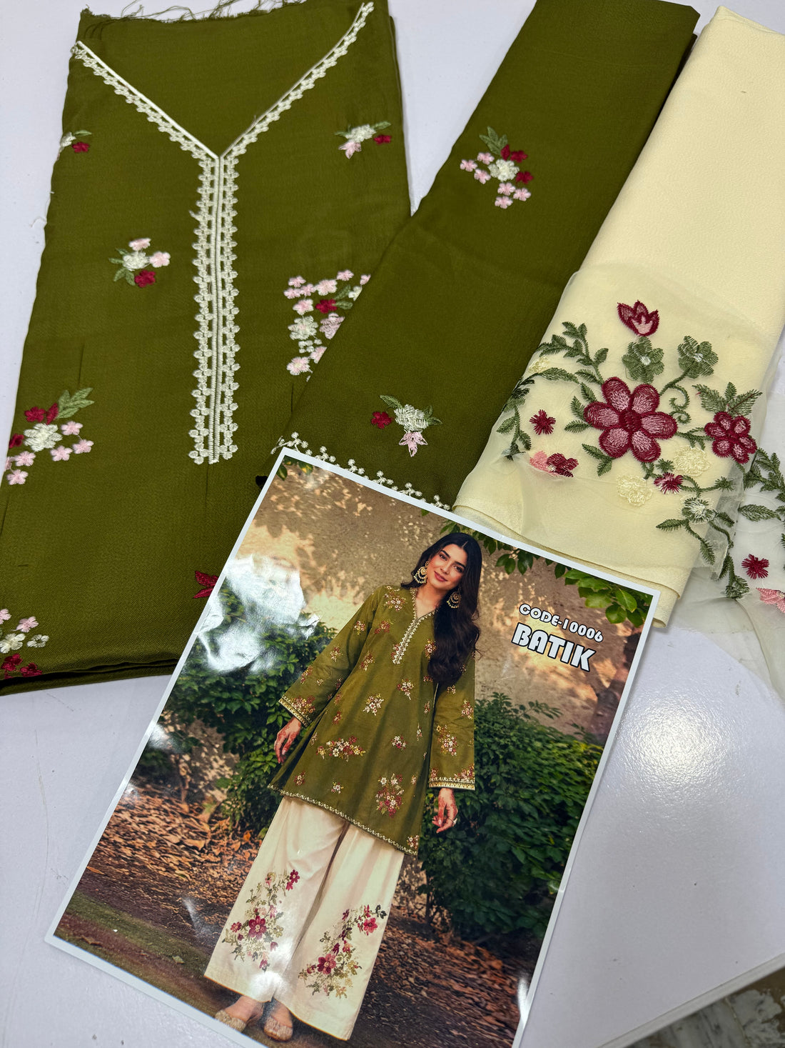 A woman stands outdoors wearing a green embroidered kurta with matching wide-leg cream pants featuring floral embroidery. The background shows green bushes and a textured wall.
