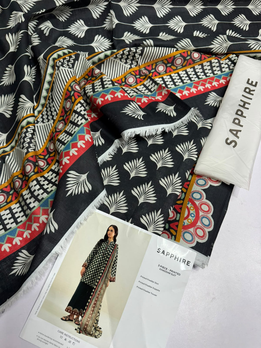 A model wearing a three-piece printed khaddar suit from Sapphire, featuring a black and beige patterned shirt, black trousers, and a matching dupatta with ornate gold and red borders, presented on a white catalog page with care instructions and product details.