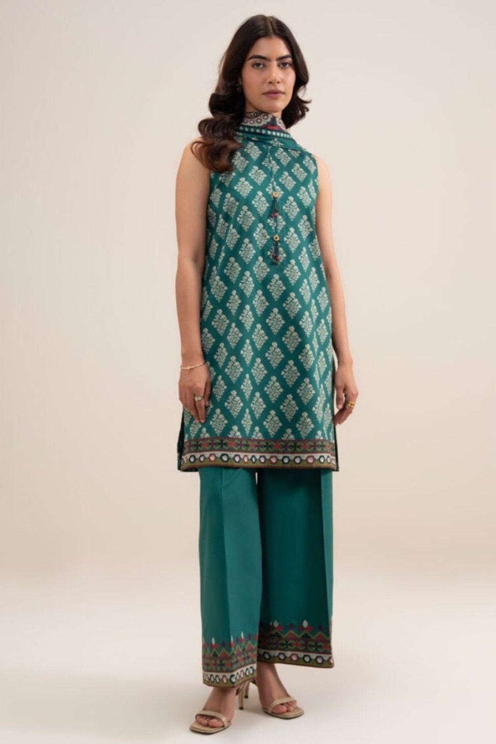 A model wears a three-piece printed khaddar suit from Sapphire, featuring a sleeveless kurta in teal and gold pattern, paired with matching teal trousers and a dupatta. The image is a product information card with care instructions, SKU number, and brand logo on a white background.
