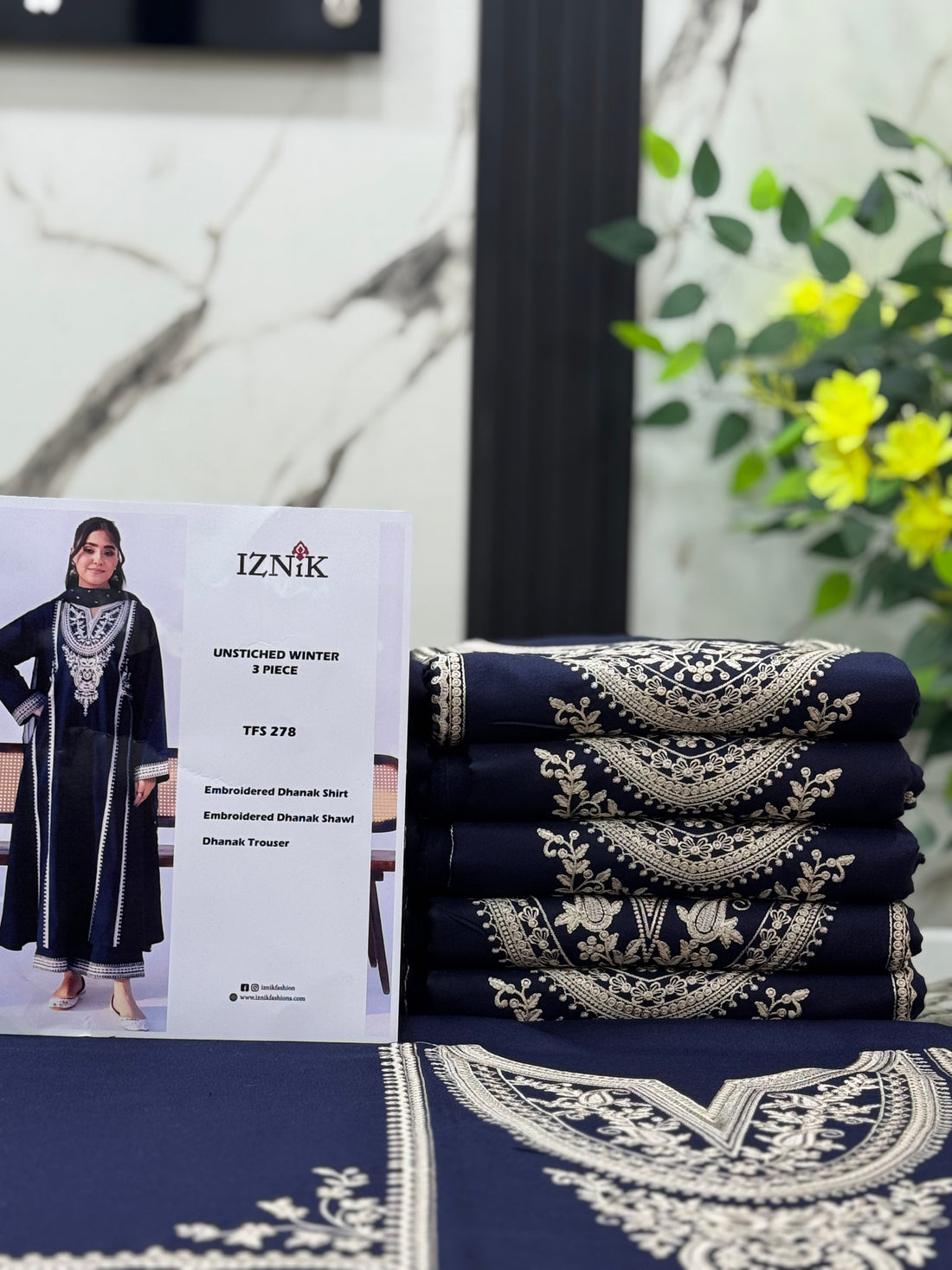 A woman modeling a navy blue long kurta with intricate silver embroidery on the chest and borders, standing in front of a wooden bench against a white background.