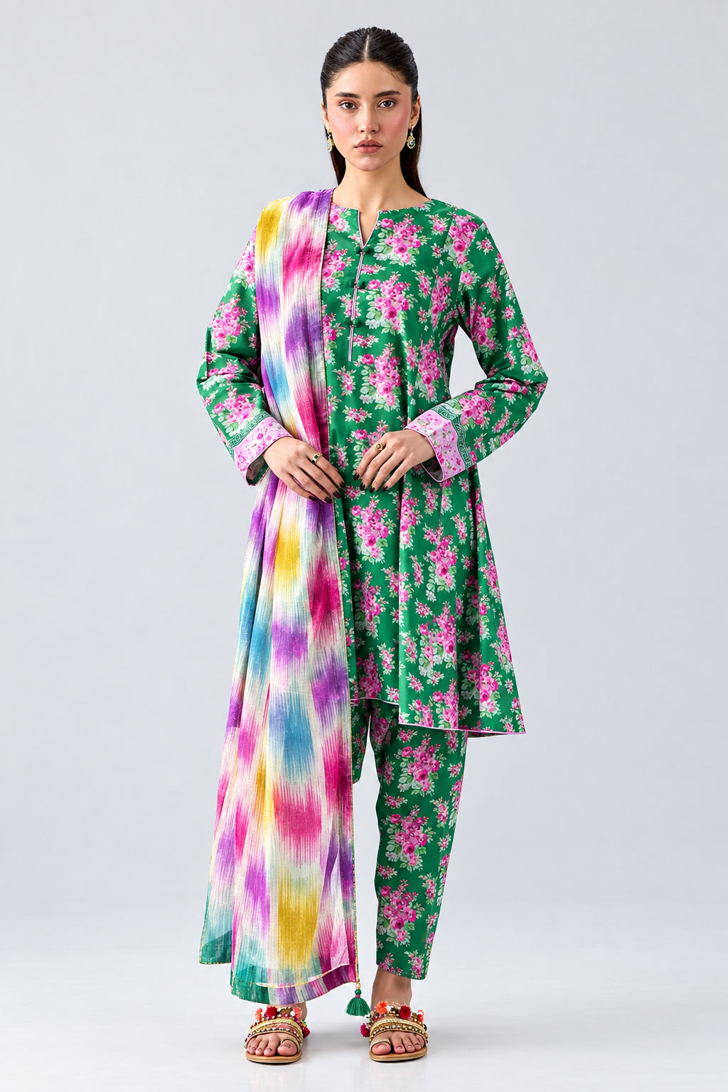 A model wearing a three-piece floral printed kurta set in green, with a matching tie-dye dupatta, on a light gray background. The image includes product details, a QR code, and care instructions under the brand name 'nishat unstitched'.
