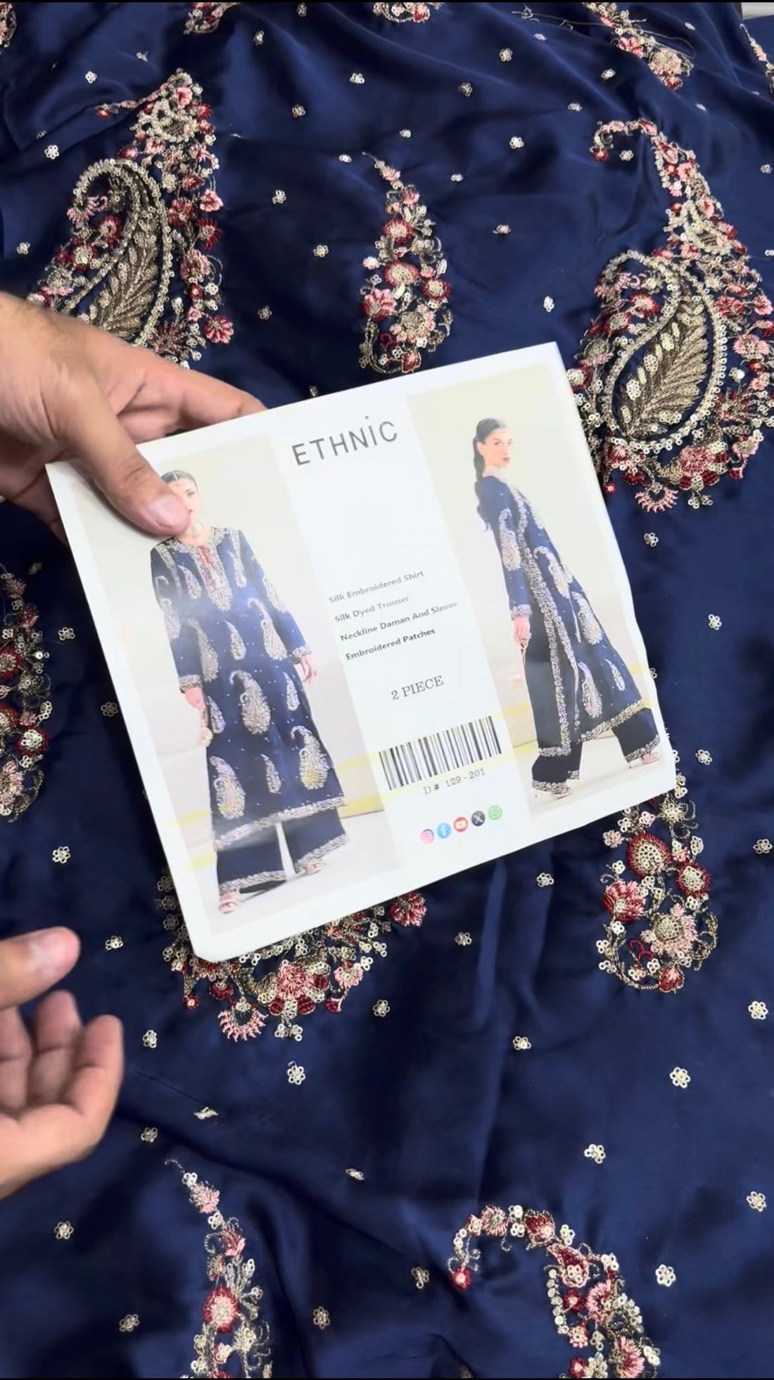 A model wearing a navy blue ethnic two-piece suit with intricate gold and silver paisley embroidery, shown in front and side views on a light beige background. The image includes text overlays describing the product as a 'Silk Embroidered Shirt' and 'Silk Dyed Trouser' with a central white panel labeled 'ETHNIC'.