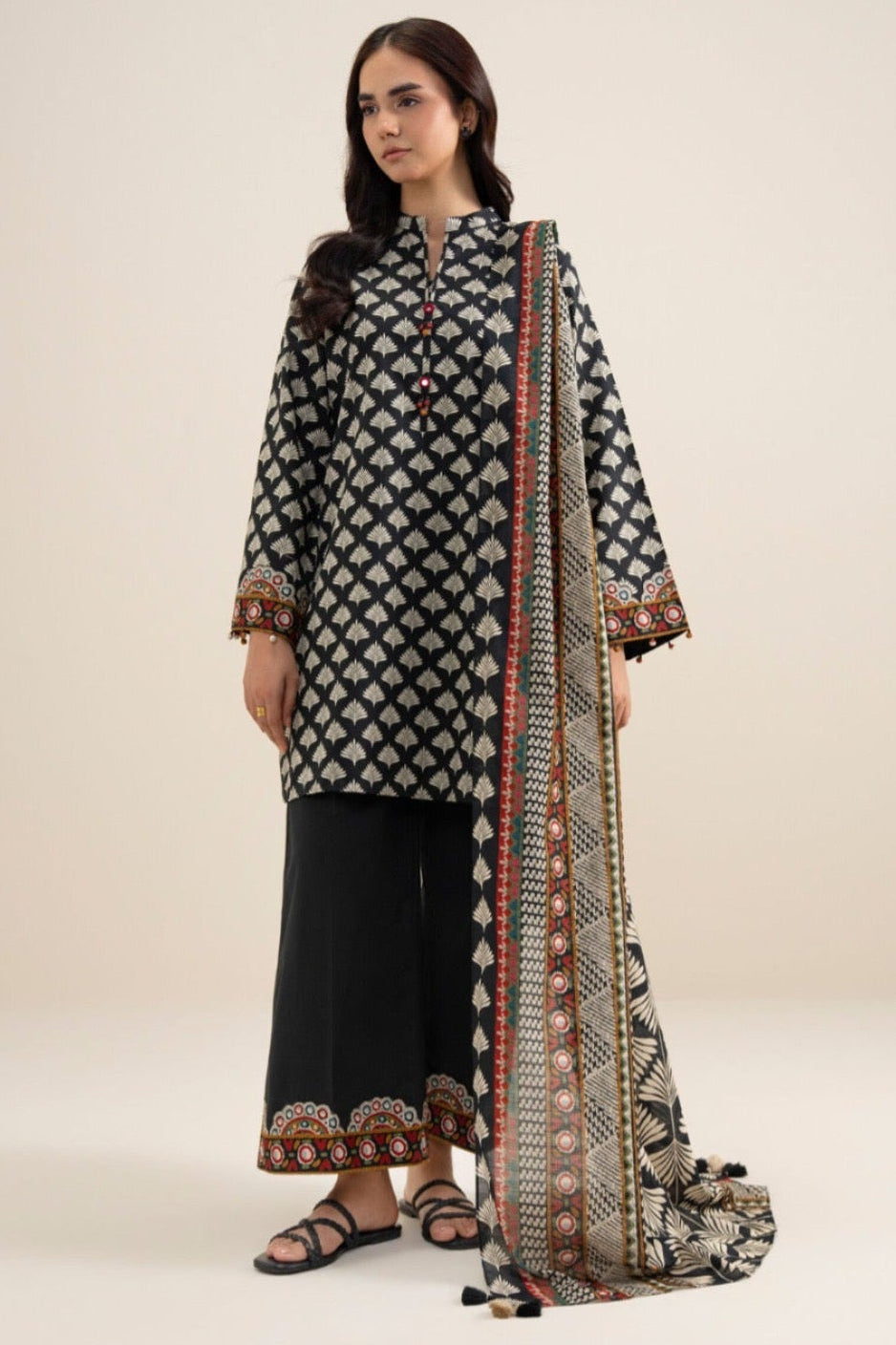 A model wearing a three-piece printed khaddar suit from Sapphire, featuring a black and beige patterned shirt, black trousers, and a matching dupatta with ornate gold and red borders, presented on a white catalog page with care instructions and product details.