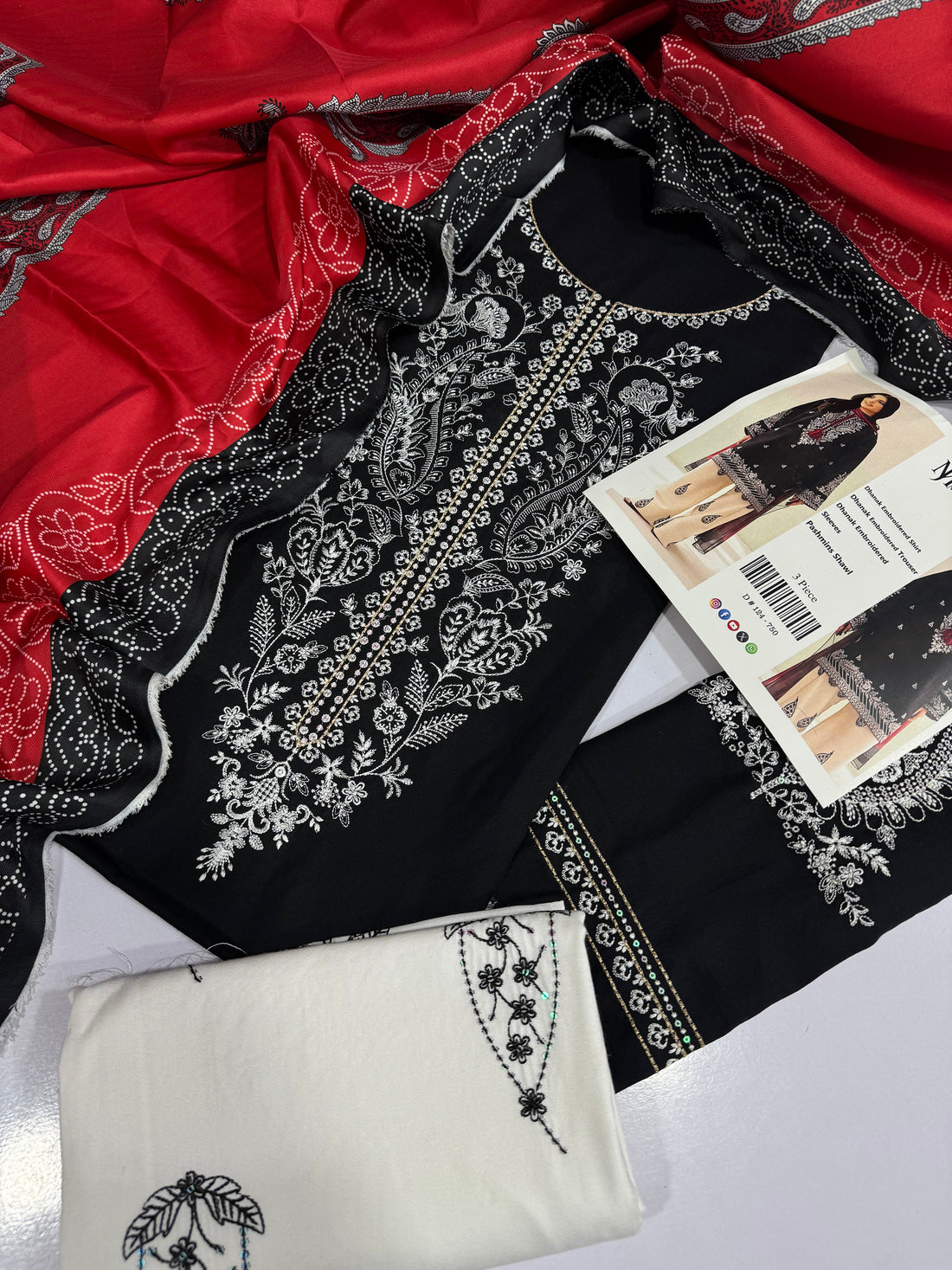 A 3-piece women's ethnic wear set from Mera Iora, featuring a black Dhanak embroidered kurta with a red shawl, beige wide-leg trousers with embroidered motifs, and a matching embroidered trouser. The image is a composite showing a model wearing the outfit on the left and right, with product details and a barcode in the center.