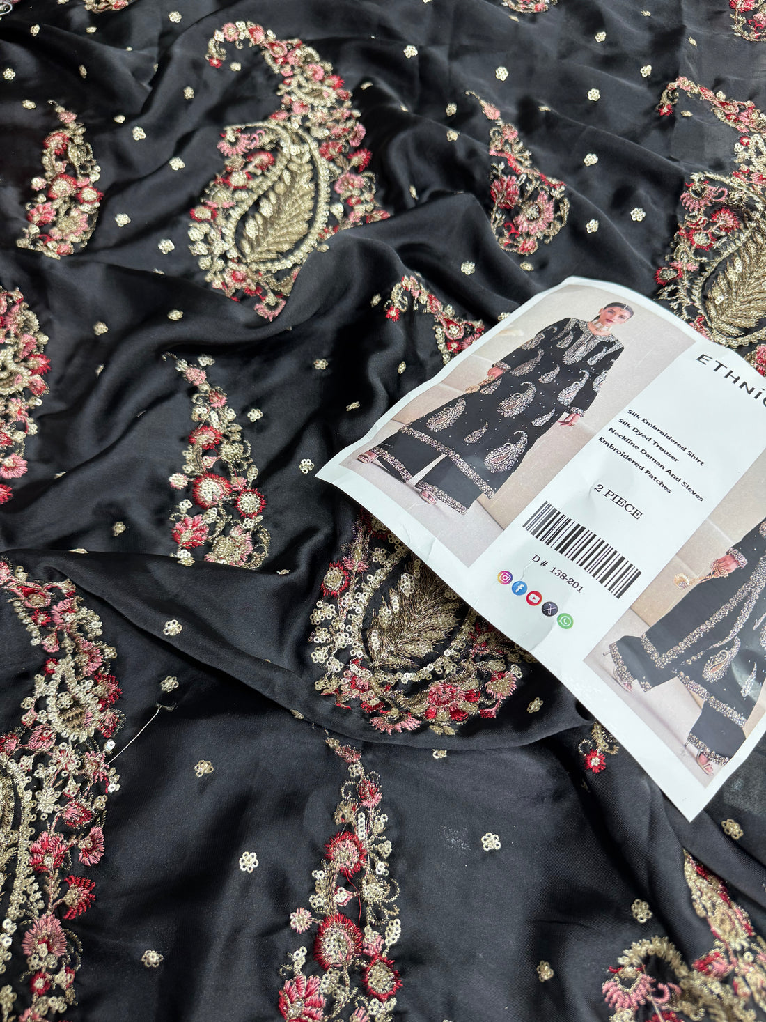 A model wearing a black ethnic two-piece suit with intricate silver and gold paisley embroidery, shown in front and side views on a beige background. The image includes text overlays describing the components of the suit and a barcode.