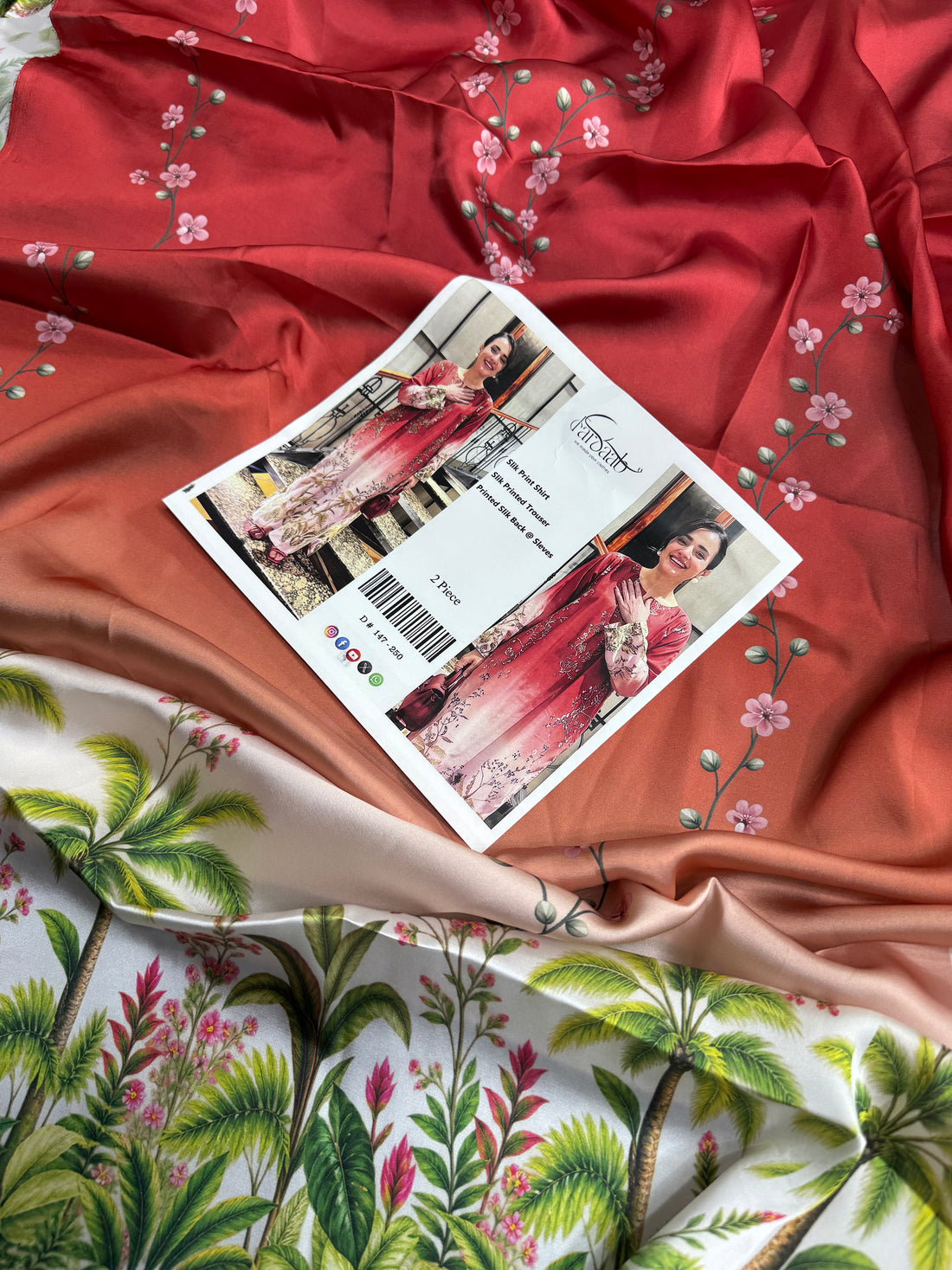 A woman modeling a two-piece silk printed outfit in an indoor staircase setting, shown in a split-screen layout with a central text panel. The outfit is a coral-orange and pink gradient tunic with beige floral patterns and matching printed trousers.