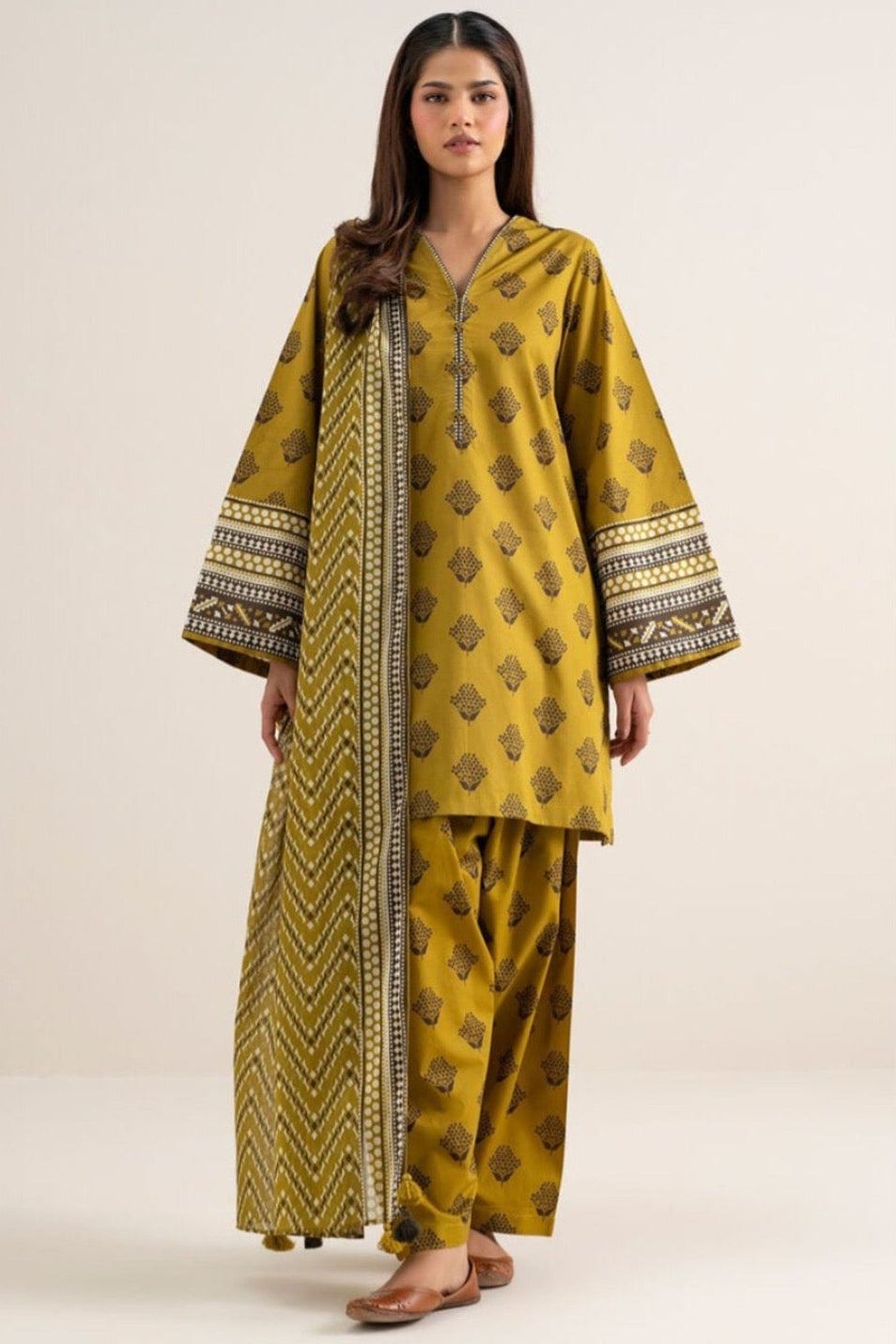 A model wearing a three-piece printed khaddar suit from the brand SAPPHIRE, featuring a mustard yellow kurta with black and gold patterns, matching trousers, and a dupatta, set against a clean white background with product details and care instructions.
