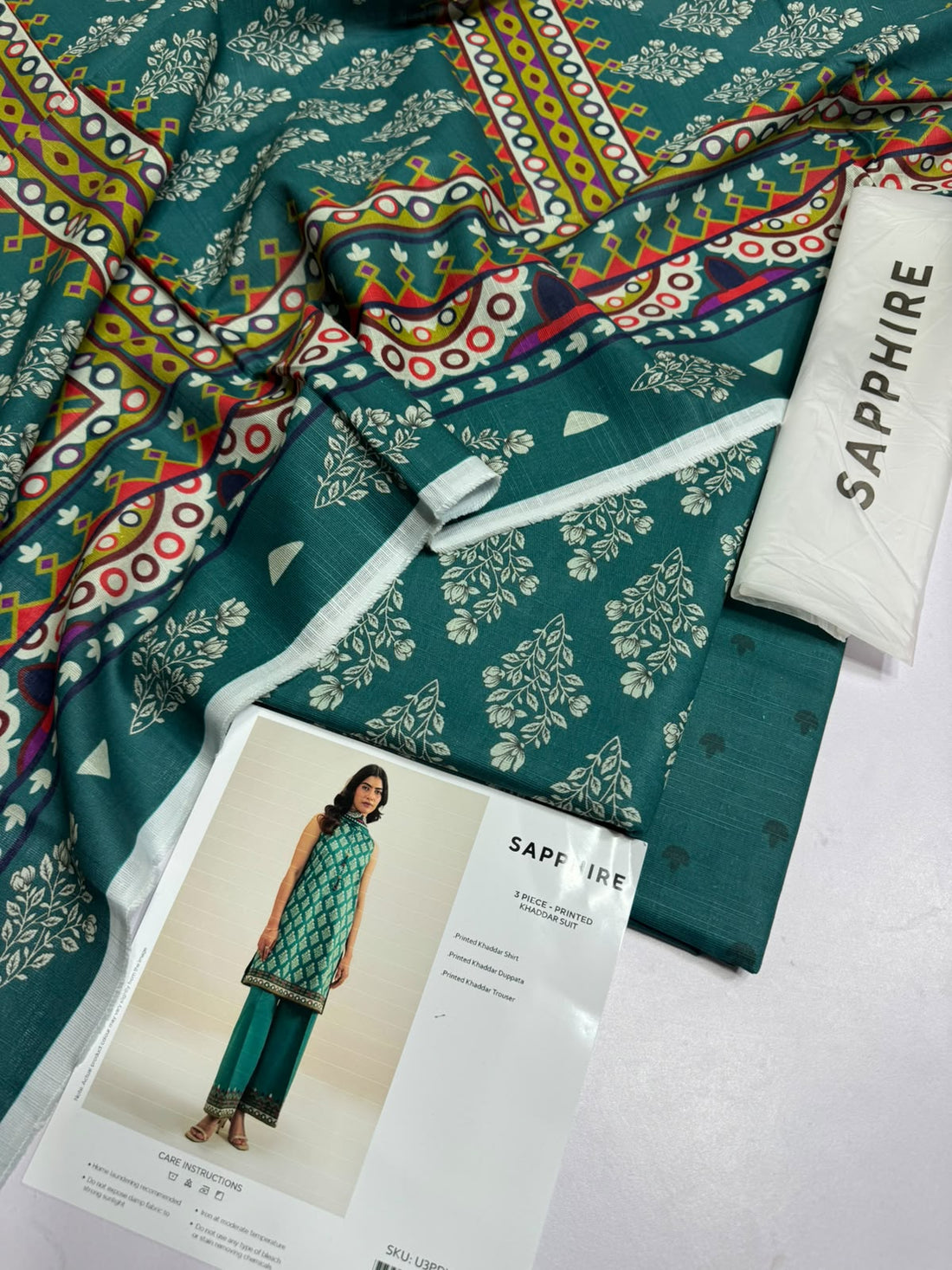 A model wears a three-piece printed khaddar suit from Sapphire, featuring a sleeveless kurta in teal and gold pattern, paired with matching teal trousers and a dupatta. The image is a product information card with care instructions, SKU number, and brand logo on a white background.