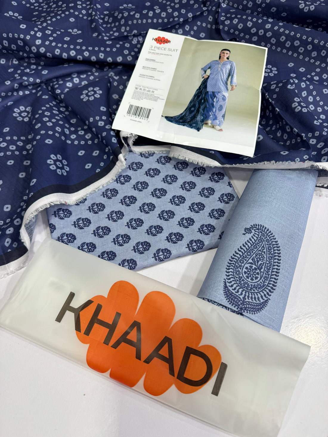 A model wearing a Khaadi 3-piece suit in lavender and navy blue, featuring a printed kurta, matching trousers, and a printed dupatta, set against a neutral studio background with product details on the left.