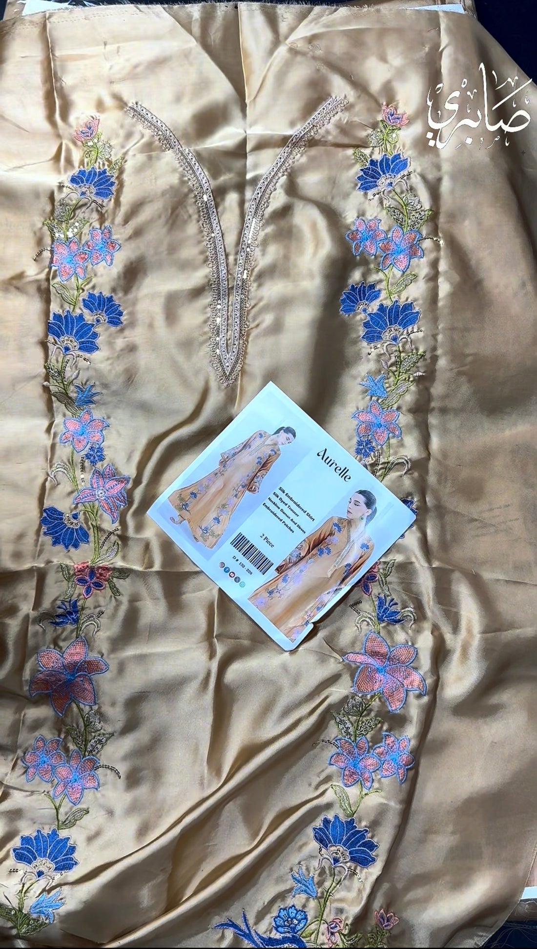 Aurelle brand catalog image showing a model wearing a beige silk embroidered shirt and trouser set. The image features a full-length view on the left, a close-up of the embroidered chest area on the right, and a central text panel detailing the product features and barcode.
