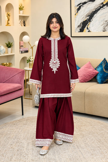 A woman modeling a deep maroon three-piece salwar kameez suit with silver mirror work embroidery on the neckline, sleeve cuffs, and pant hems, standing in a modern living room setting.