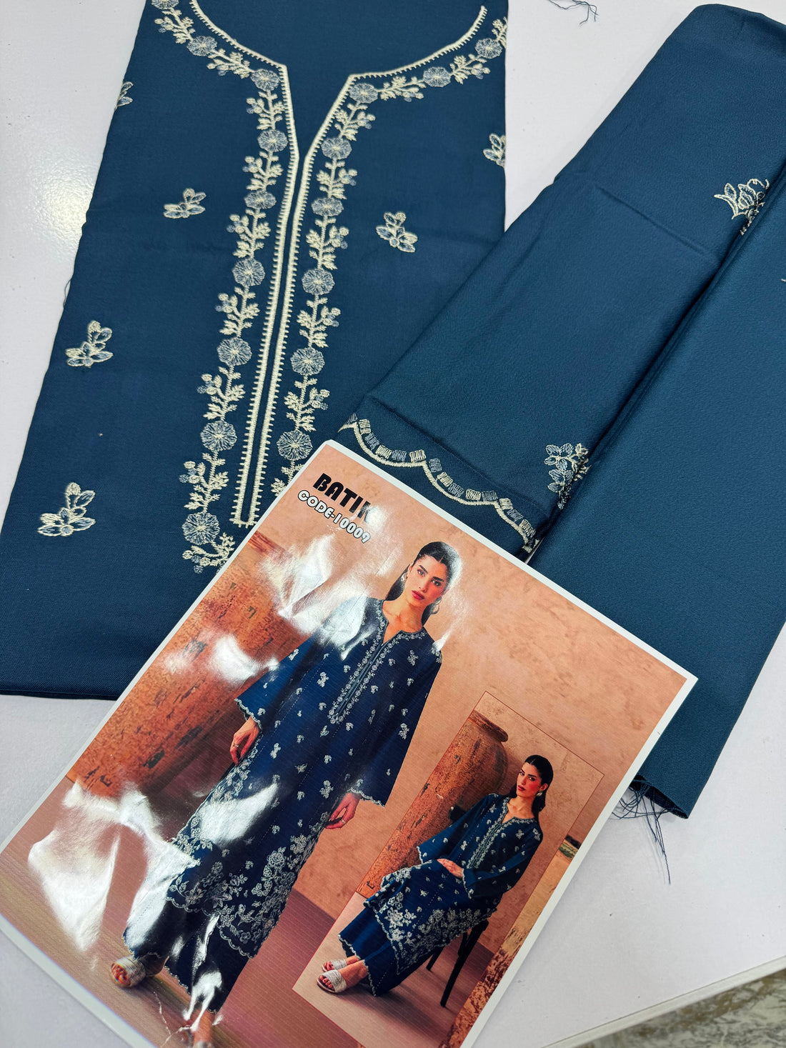 A woman models a navy blue Batik kurta set with white floral block prints and matching pants in a studio setting with rustic clay pots. An inset shows a seated view of the same outfit.