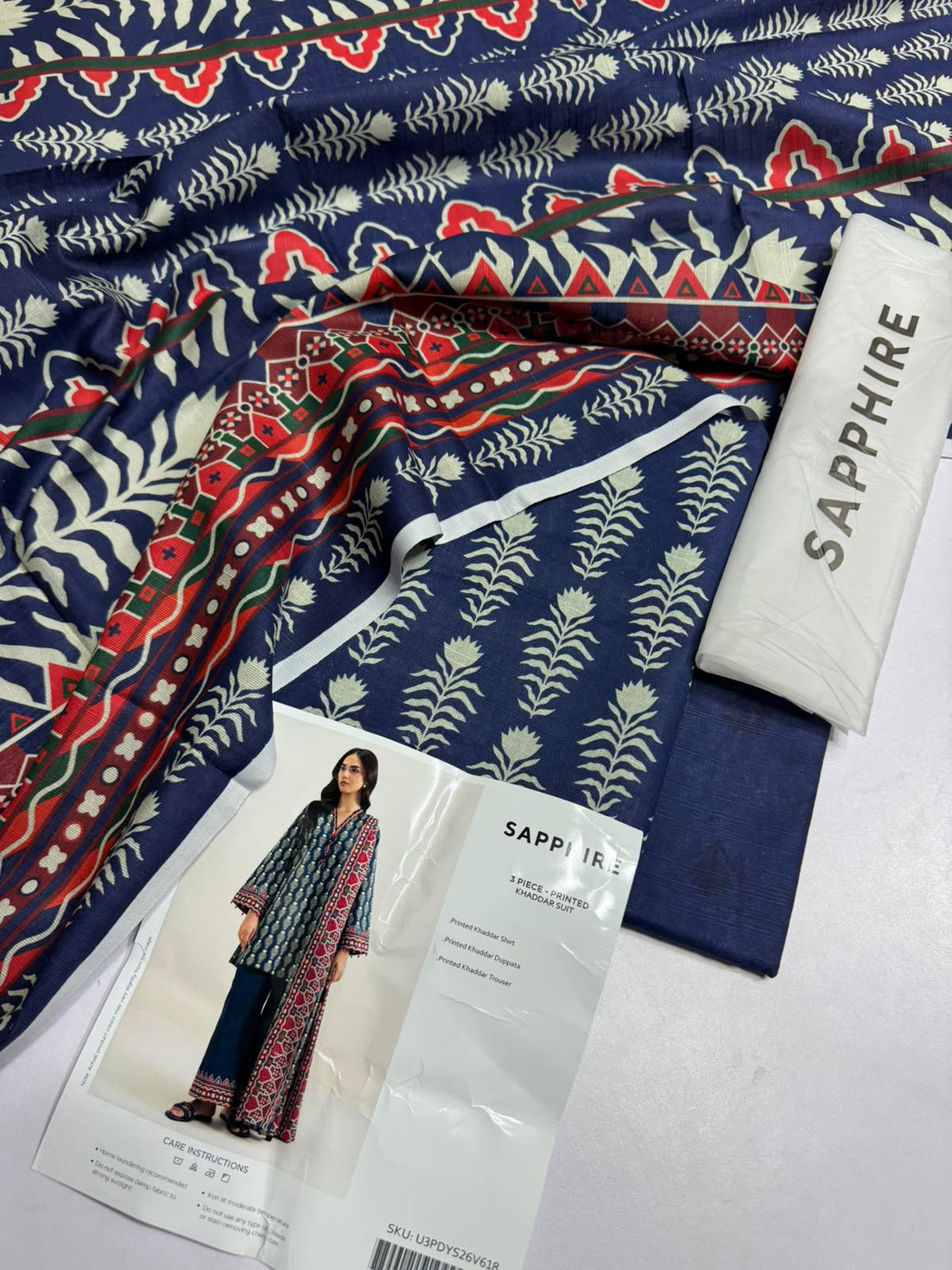 A model wearing a three-piece printed khaddar suit by Sapphire, featuring a long kurta, trousers, and a top, displayed on a clean white catalog card with product details and care instructions.