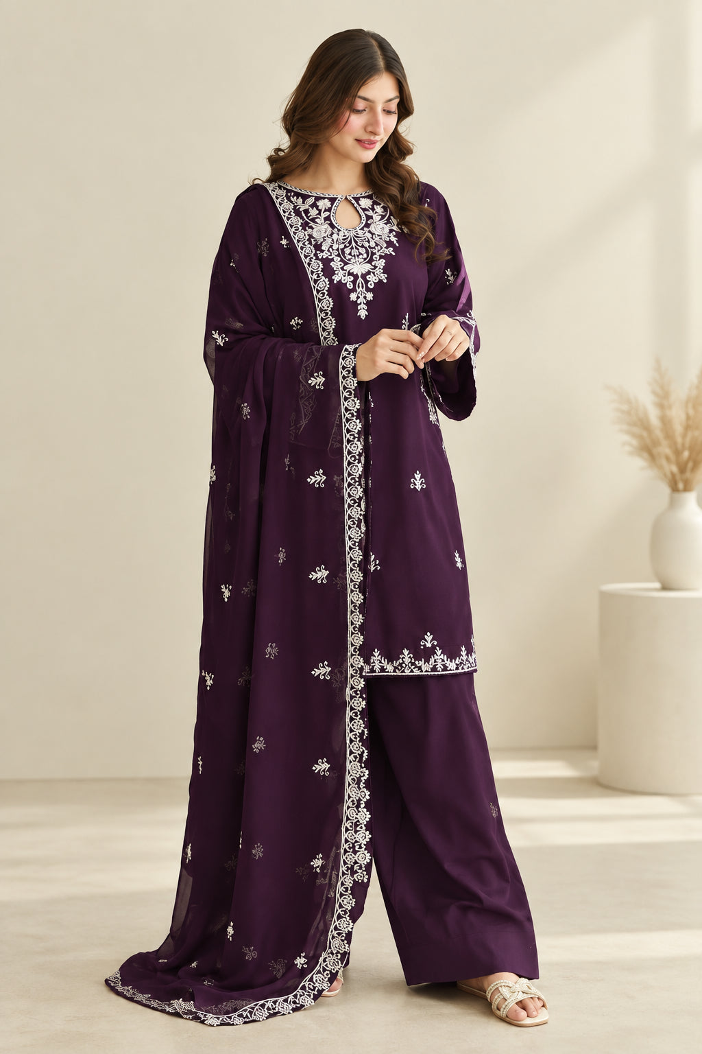 A woman models a deep purple three-piece traditional outfit with white embroidery, featuring a long kurta, matching trousers, and a draped dupatta. The image includes an inset showing the back view of the outfit and a brand logo 'LAAM' in the top right corner.