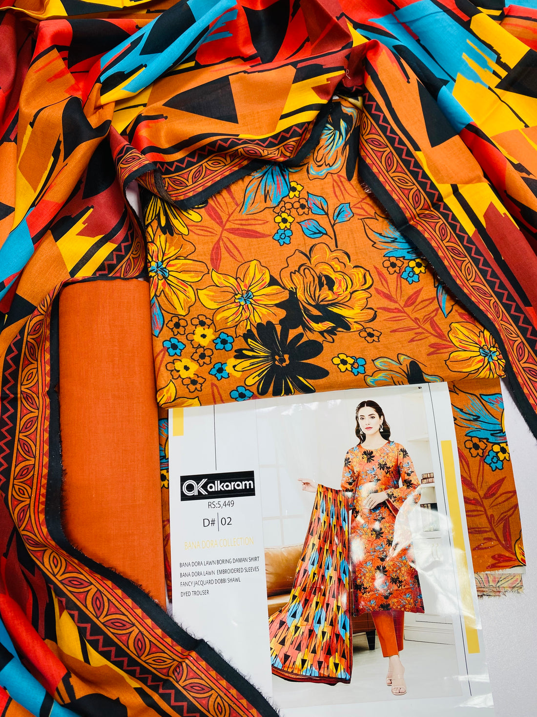 A woman modeling a Kalkaram Bana Dora Lawn/Borderline set, featuring a bright orange kurta with black floral patterns, matching orange pants, and a coordinating printed dupatta, set against an indoor home background.