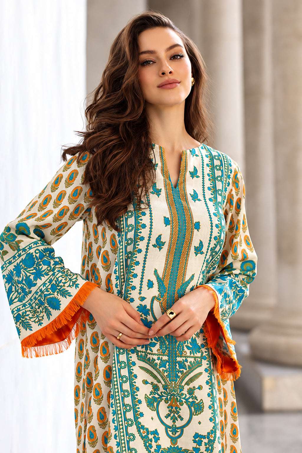 A woman wearing a cream-colored long kurta with blue and orange printed patterns, standing in front of a blurred stone column background.