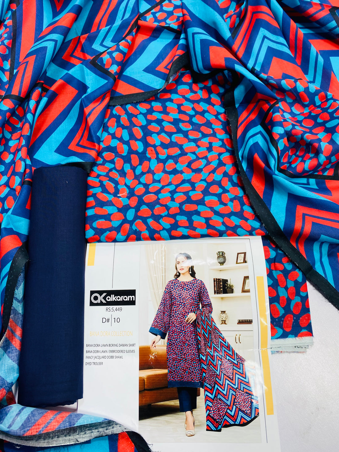 A woman modeling a Kalkaram Bana Dora collection three-piece suit. The outfit consists of a blue and red patterned kurta, dark blue pants, and a matching patterned dupatta draped over her shoulder.