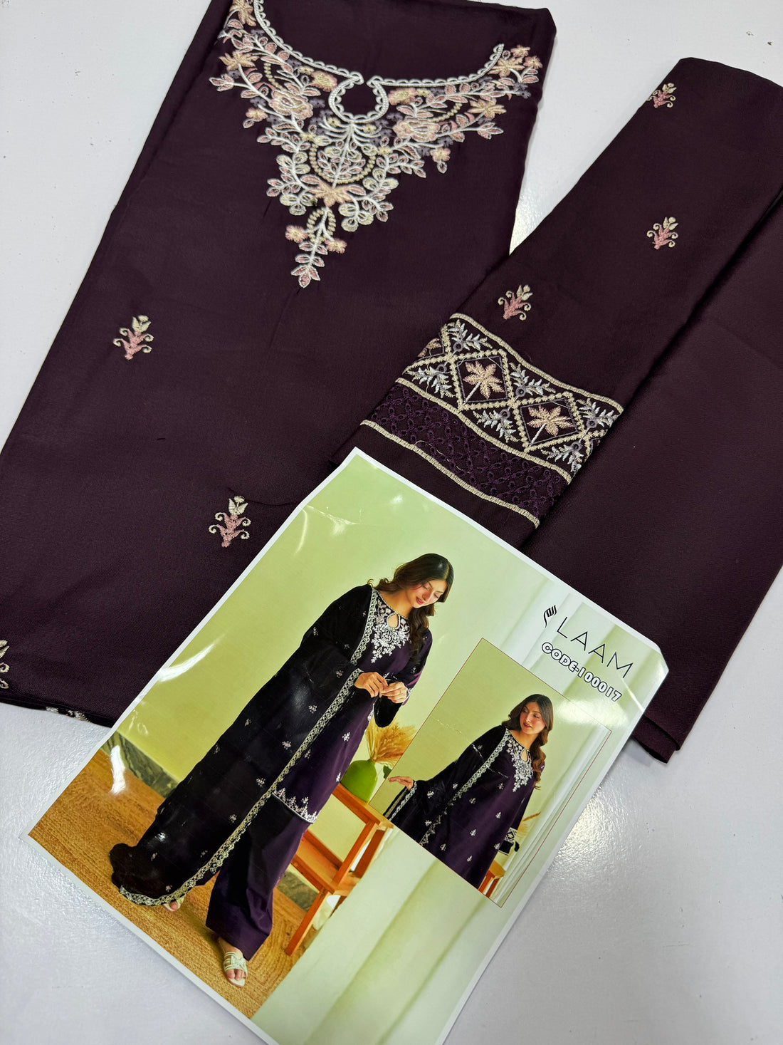 A woman models a deep purple three-piece traditional outfit with white embroidery, featuring a long kurta, matching trousers, and a draped dupatta. The image includes an inset showing the back view of the outfit and a brand logo 'LAAM' in the top right corner.