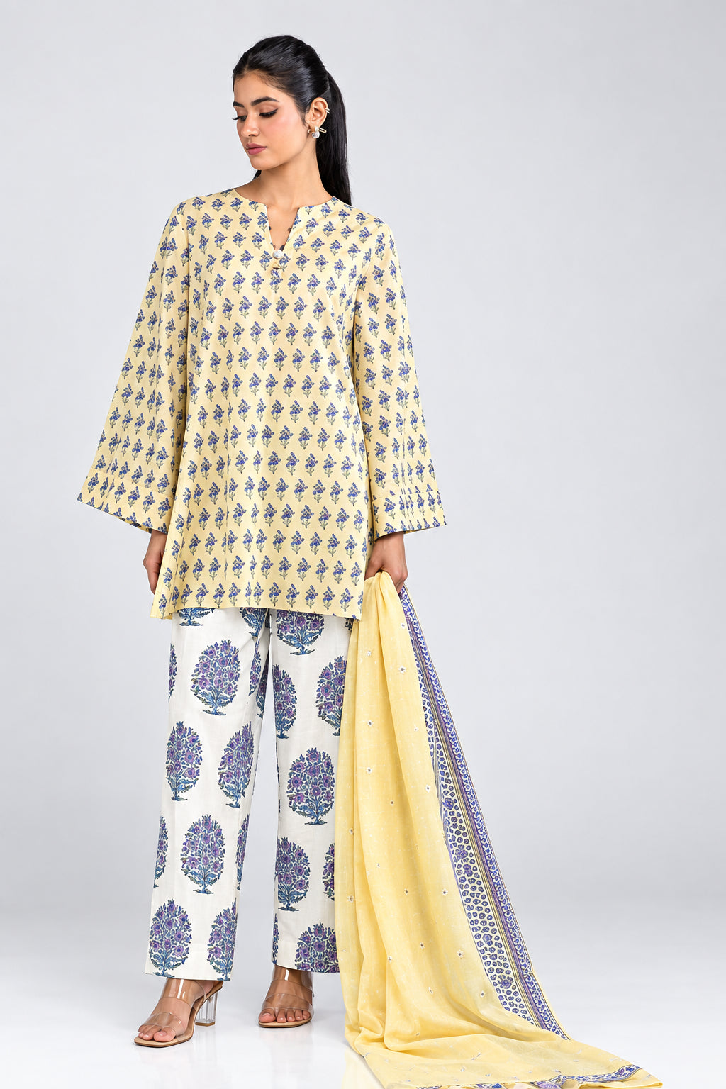 A woman models a Khaadi 3-piece suit set in a studio setting. The set includes a cream-colored printed kurta, matching printed trousers, and a light yellow dupatta with purple borders.