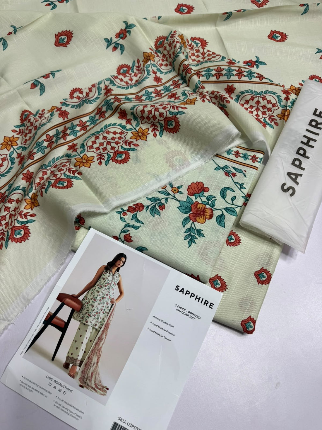 A model wears a three-piece printed khaddar suit from Sapphire, featuring a sleeveless kurta, matching trousers, and a dupatta. The set has a cream base with a vibrant multicolor floral print.