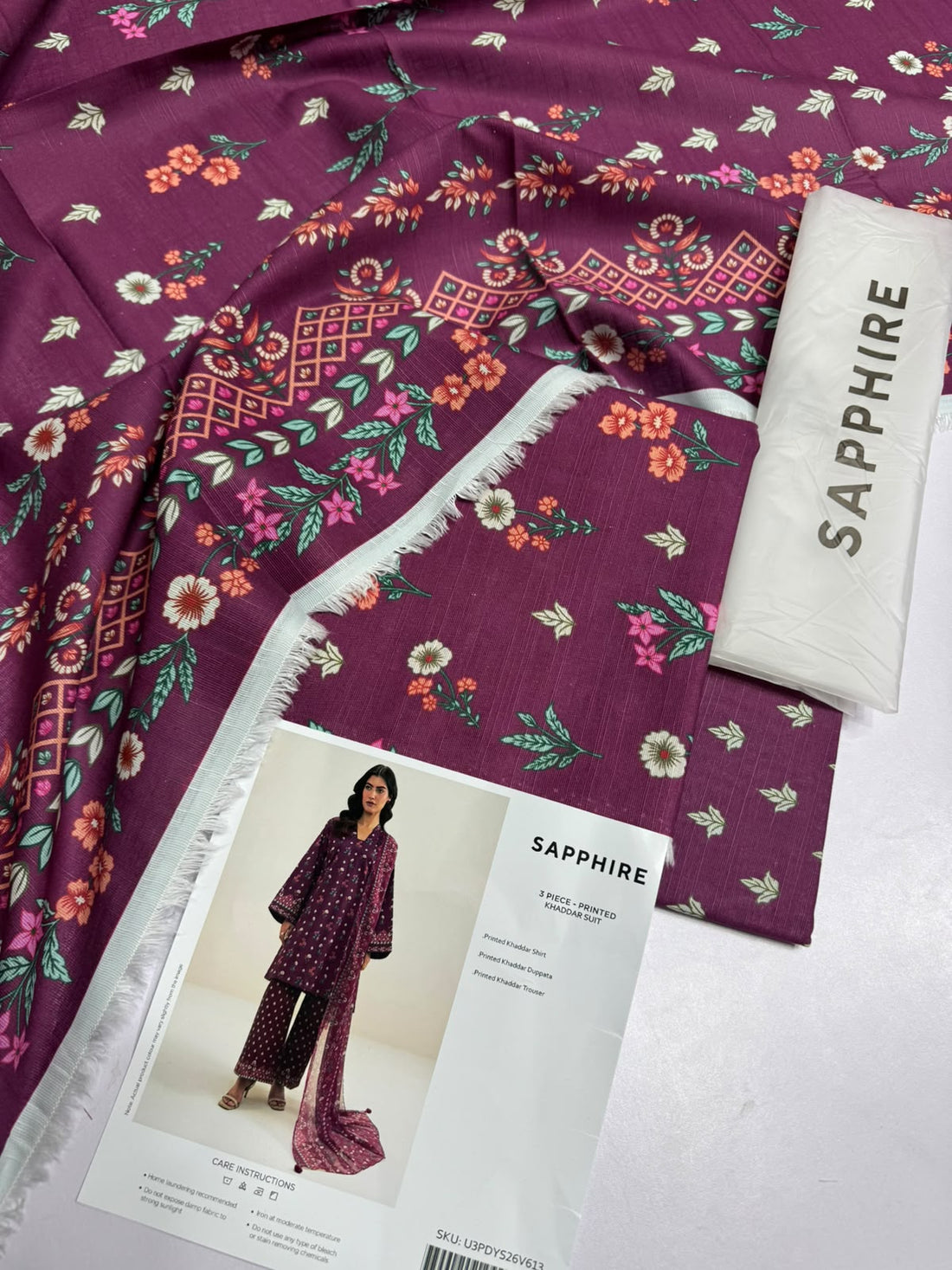 A model wearing a three-piece printed khaddar suit from Sapphire, featuring a dusty purple kurta, matching trousers, and a dupatta with small yellow and white floral patterns, set against a plain light beige background with product details on the right.