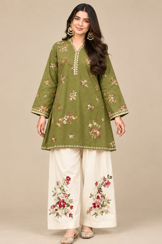 A woman stands outdoors wearing a green embroidered kurta with matching wide-leg cream pants featuring floral embroidery. The background shows green bushes and a textured wall.
