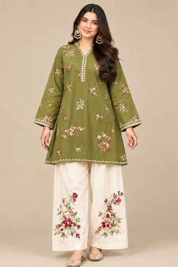 A woman stands outdoors wearing a green embroidered kurta with matching wide-leg cream pants featuring floral embroidery. The background shows green bushes and a textured wall.