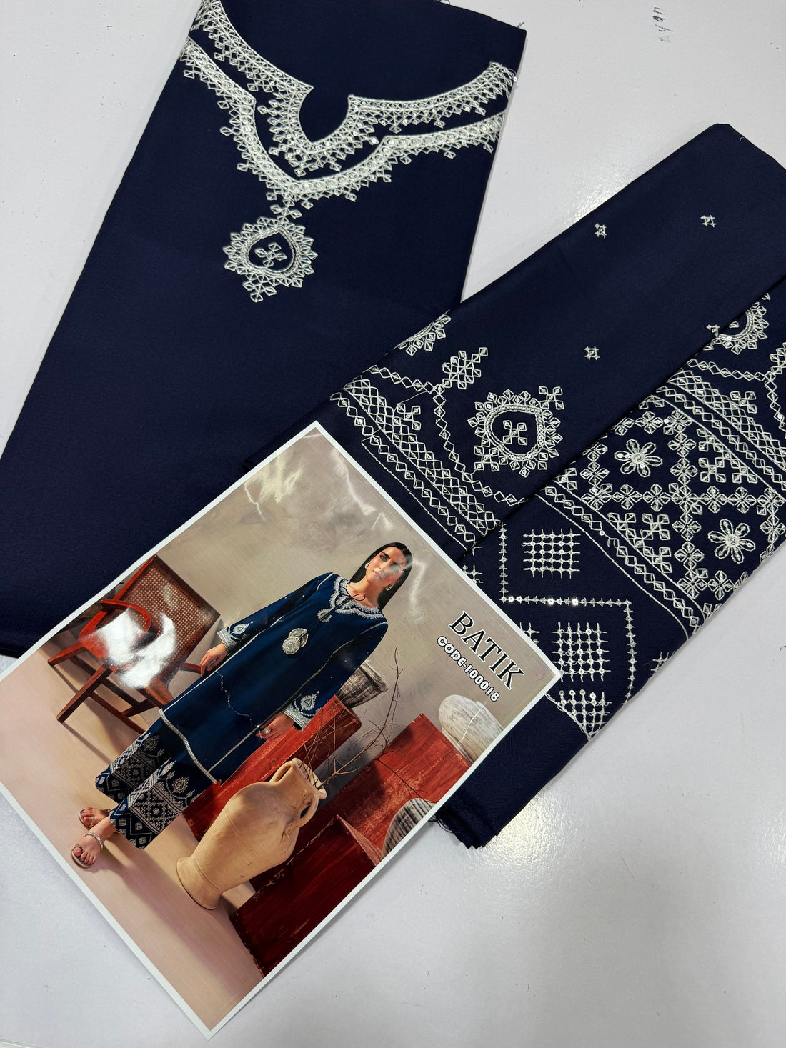 A woman models a navy blue batik kurta and trouser set with intricate white and silver embroidery, standing in a styled studio setting with wooden furniture and ceramic vases. The text 'BATIK CODE:100018' is visible in the top right corner.