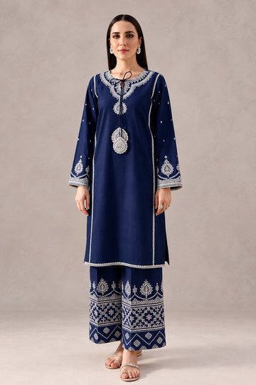 A woman models a navy blue batik kurta and trouser set with intricate white and silver embroidery, standing in a styled studio setting with wooden furniture and ceramic vases. The text 'BATIK CODE:100018' is visible in the top right corner.
